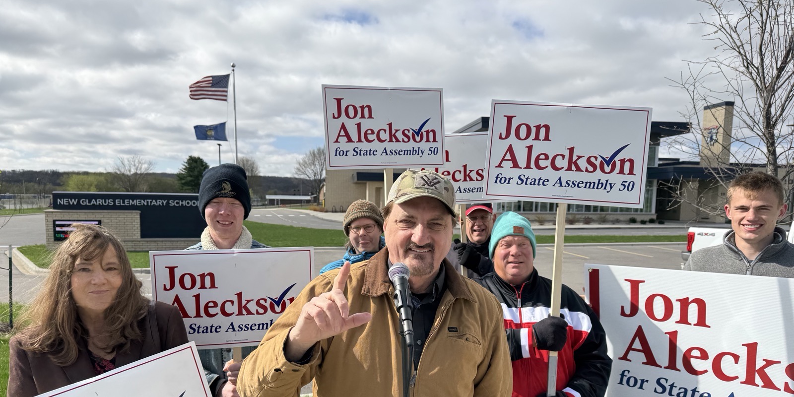 Jon Aleckson speaking with supporters holding campaign signs in front of New Glarus Elementary School